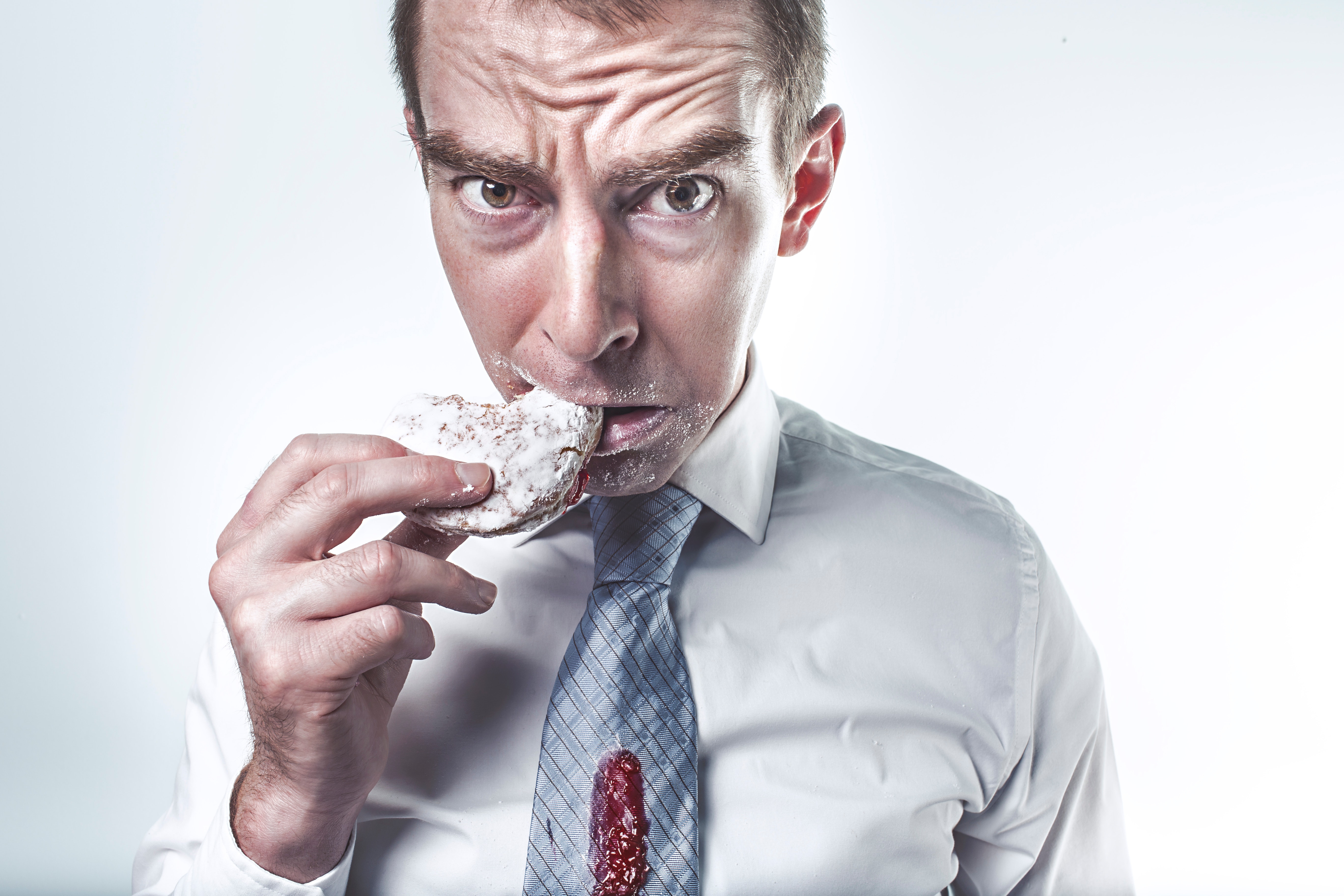 Picture shows a hungry/angry white man wearing a shirt and tie and eating a powdered donut.
