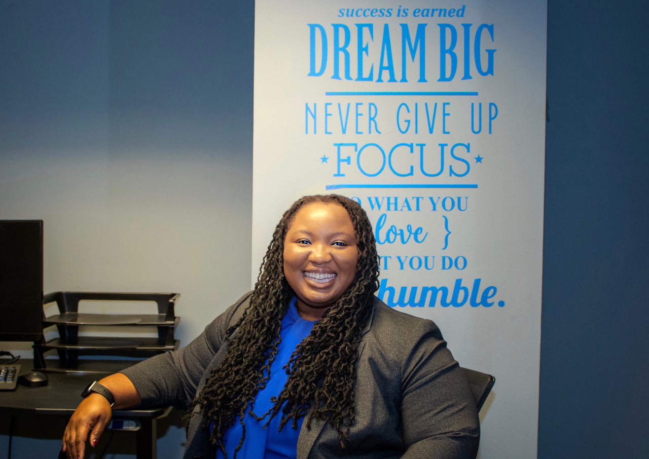 Shauna Cooper and her team at the Strengths, Assets and Resilience Lab explore the ways in which families, schools and communities influence positive development among African-American adolescents and youth. (photo by Kristen Chavez), Cooper is sitting in front of a door that has words in blue on it like: "Dream Big. Never Give Up. Focus. Success is Earned. etc."