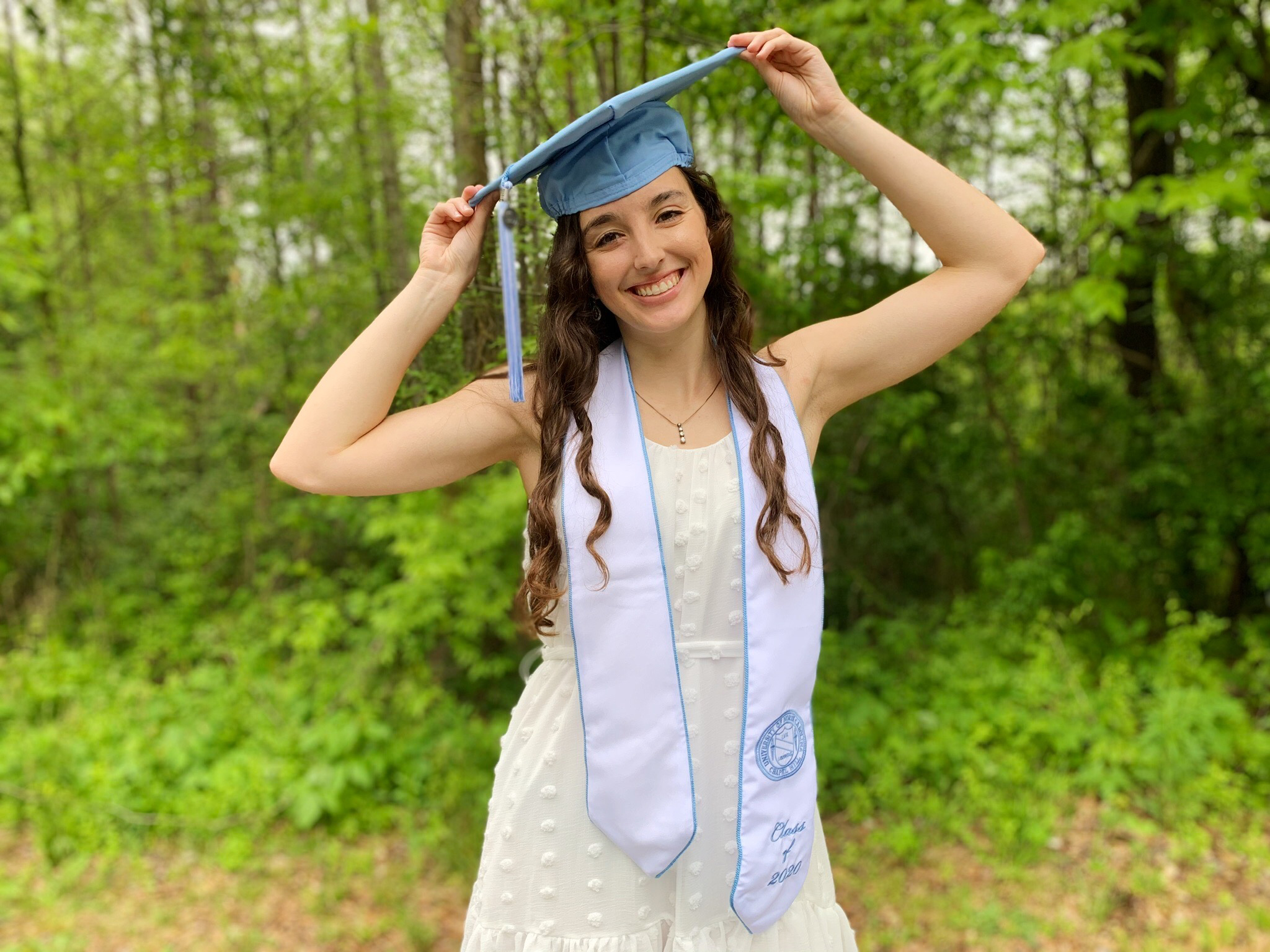 Lauren Revis stands in a yard with her graduation cap on in a white dress.
