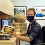 Brett Harris, masked, sits at a desk and holds an open book. A globe is in the background with the sign Center for European Studies.