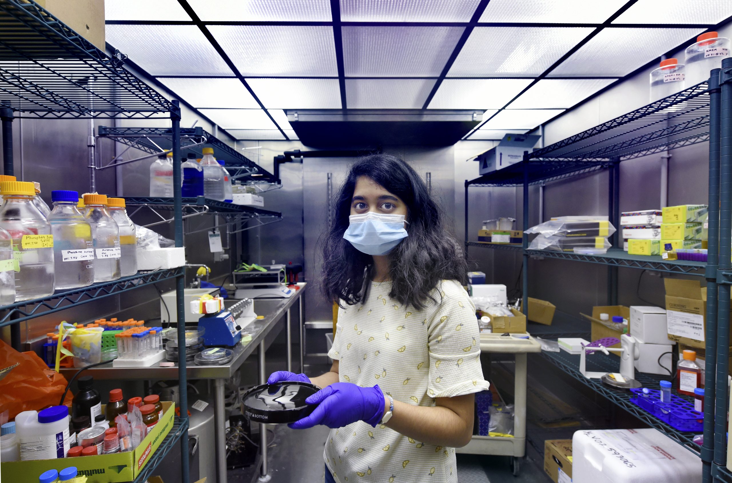 Sriya Kongala stands masked in a lab wearing purple gloves with lab equipment behind her. 