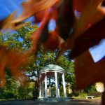 A view of the Old Well from across Cameron Avenue peeking through the leaves of an orange-brown tree. (photo by Donn Young)