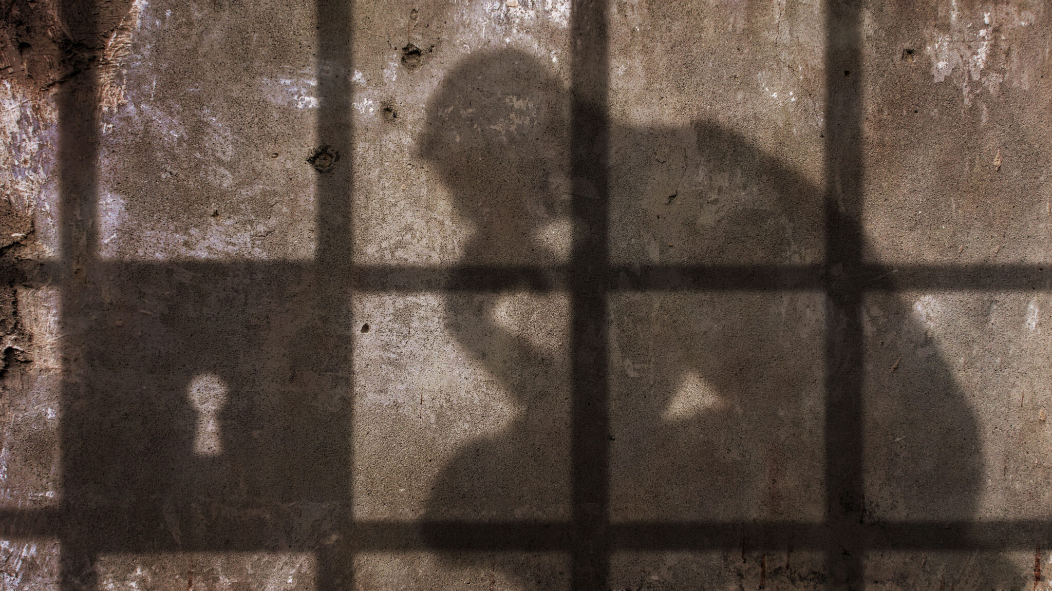 A man's shadow (sitting down, leaning forward with hands on knees) is seen behind the bars of a prison cell.