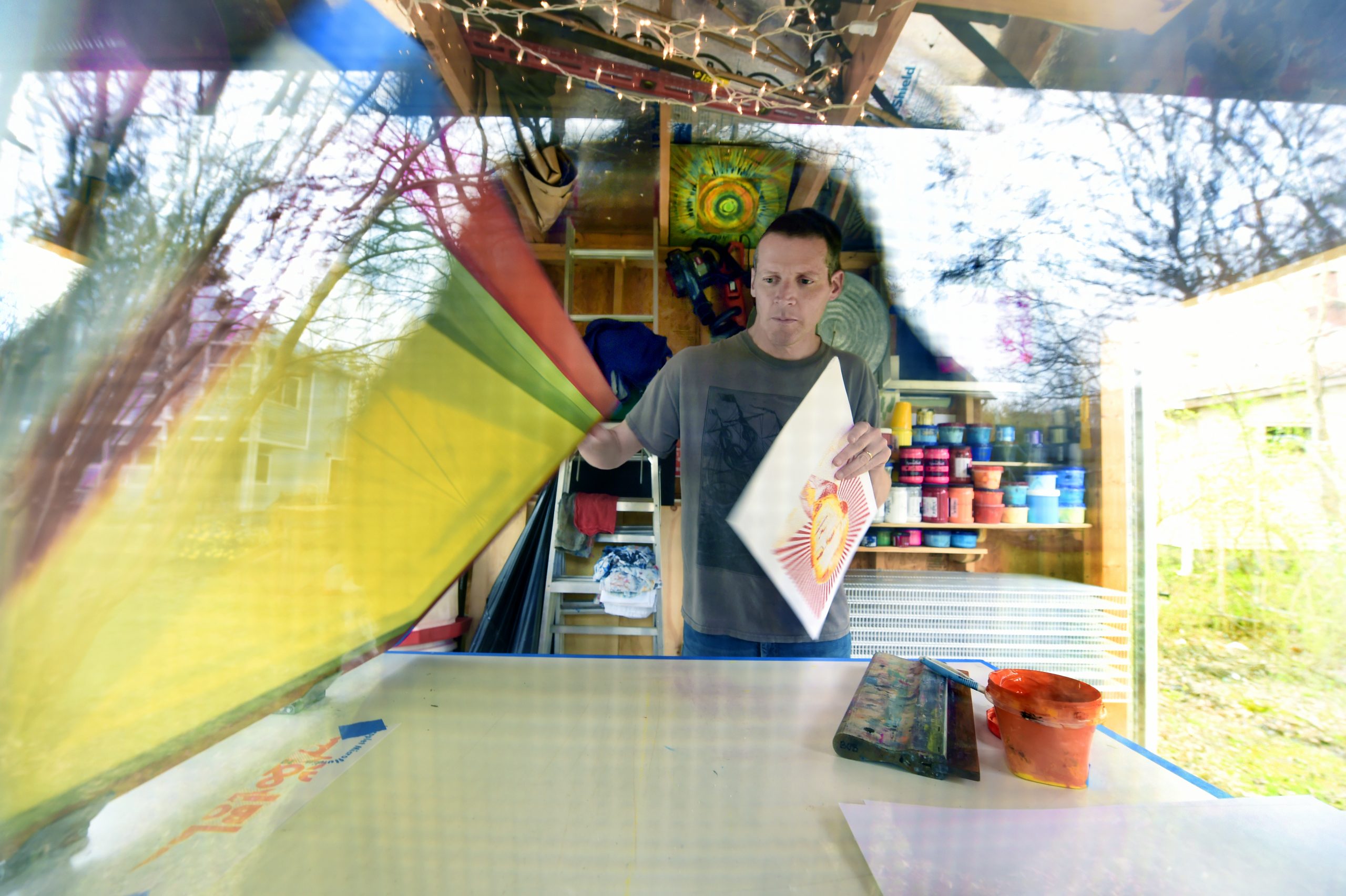Bob Goldstein lifts the lid on a screen printer as he he is surrounded by bright colors in his shed where he does his art.