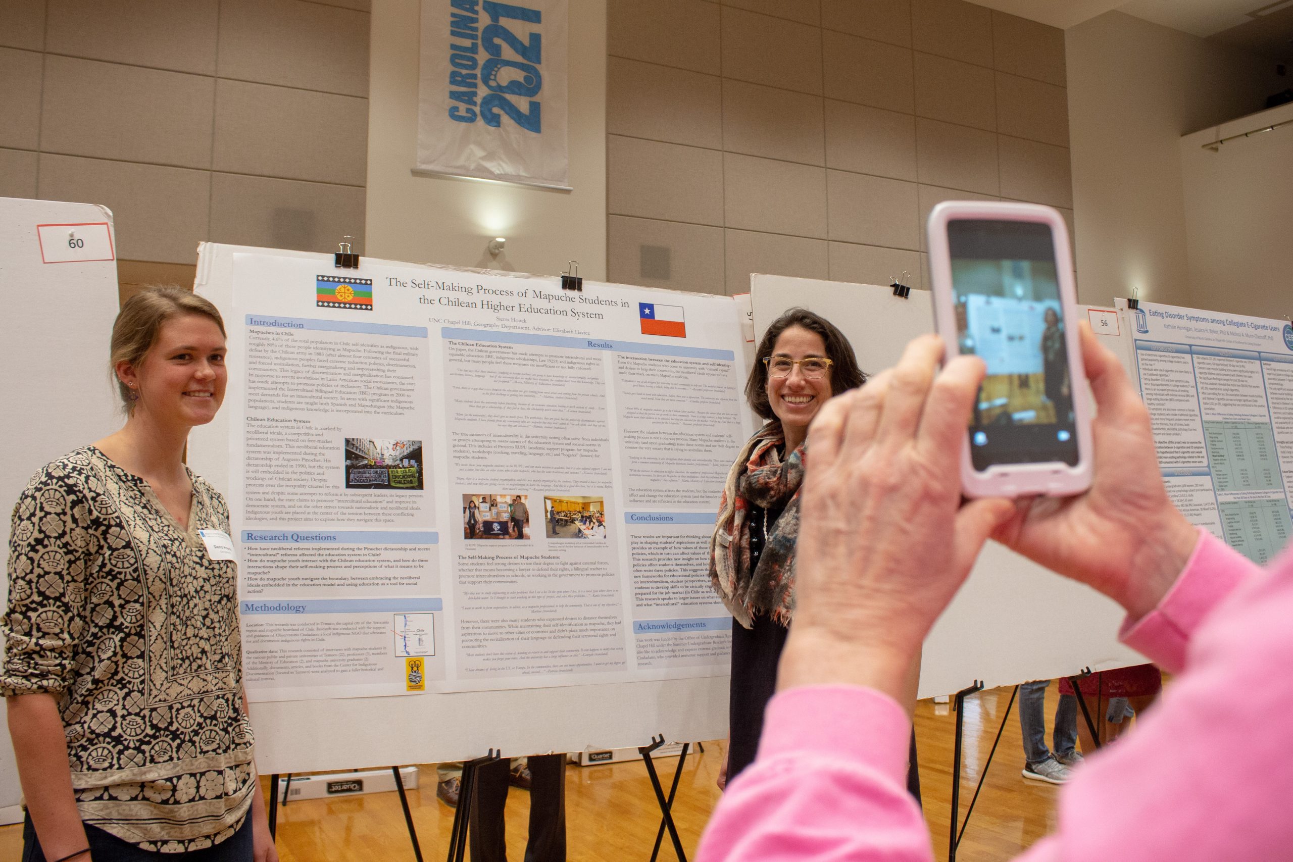 Sierra Houck and her advisor Elizabeth Havice pose for a photo taken by Houck's parents. Behind them is Houck's research poster, "The Self-Making Process of  Mapuche Students in the Chilean Higher Education System"
