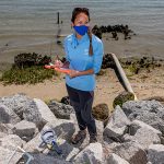 Stacy Zhang stands on a beach in Morehead City with a mask on conducting research.