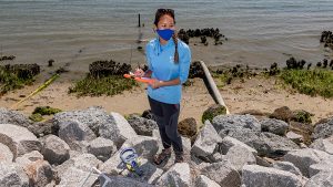 Stacy Zhang stands on a beach in Morehead City with a mask on conducting research.