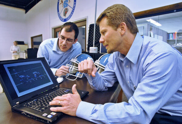 Jason Mihalik and Kevin Guskiewicz look at a computer screen. Mihalik holds a football helmet.