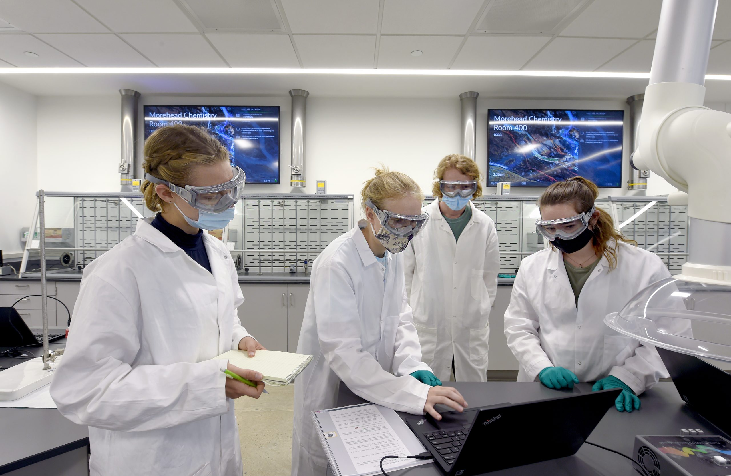 Students in lab coats and masks stand around a monitor in the lab.