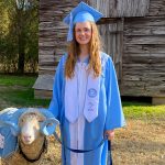 Hannah King stands in cap and gown beside Rameses XXII outside on a farm.