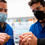 Erik Alexanian and Frank Leibfarth (left to right) dressed in lab gear and masks pull on a piece of plastic.