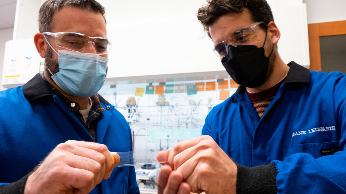 Erik Alexanian and Frank Leibfarth (left to right) dressed in lab gear and masks pull on a piece of plastic.