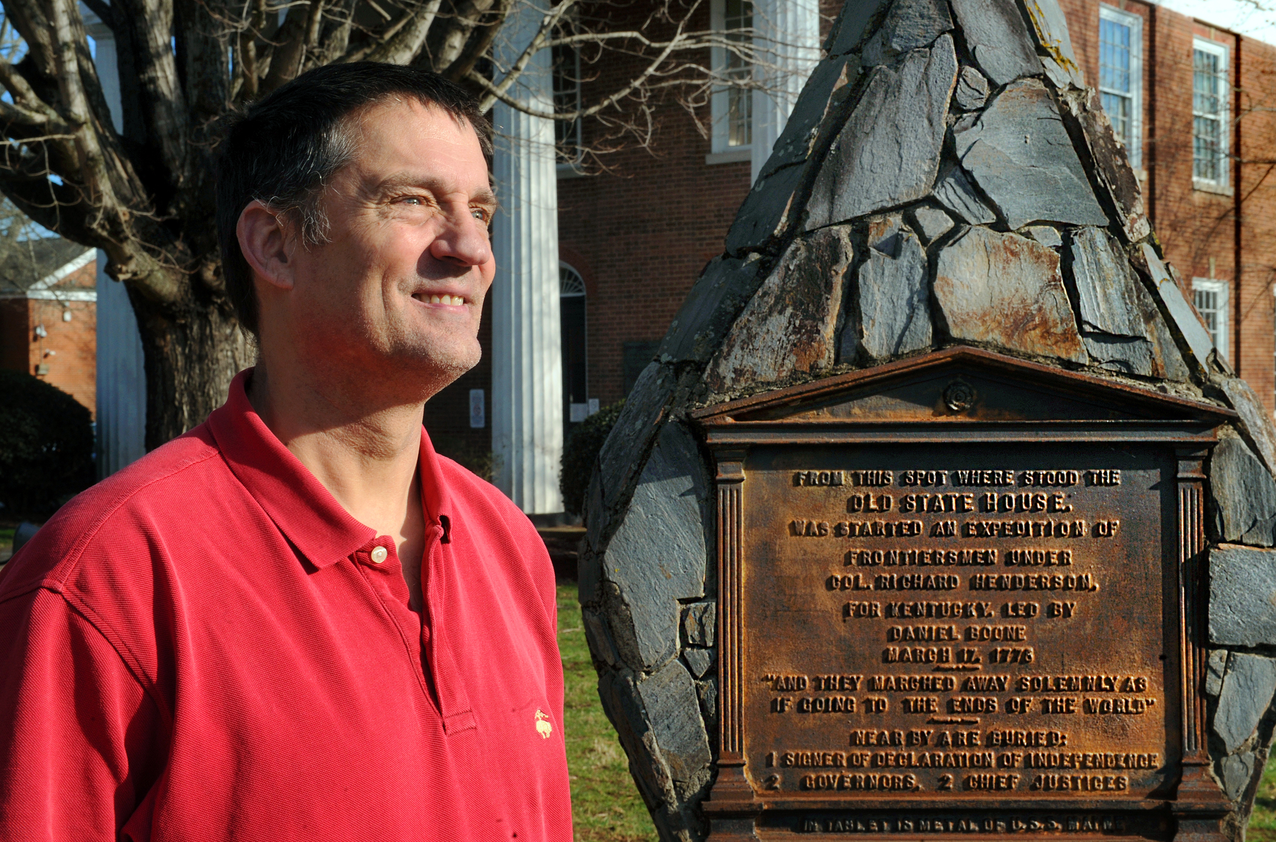 Fitz Brundage, wearing a red shirt, looks off to the right as he stands beside a monument in downtown Hillsborough.