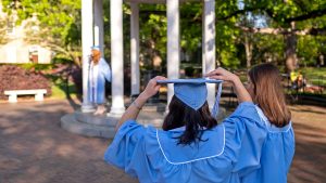Two female graduates in caps and gowns stand with their backs to the camera facing the Old Well.