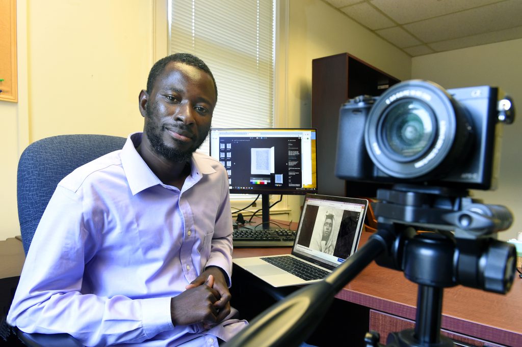 Samba Camara sits in his office with a camera in the foreground and his computer in the background.