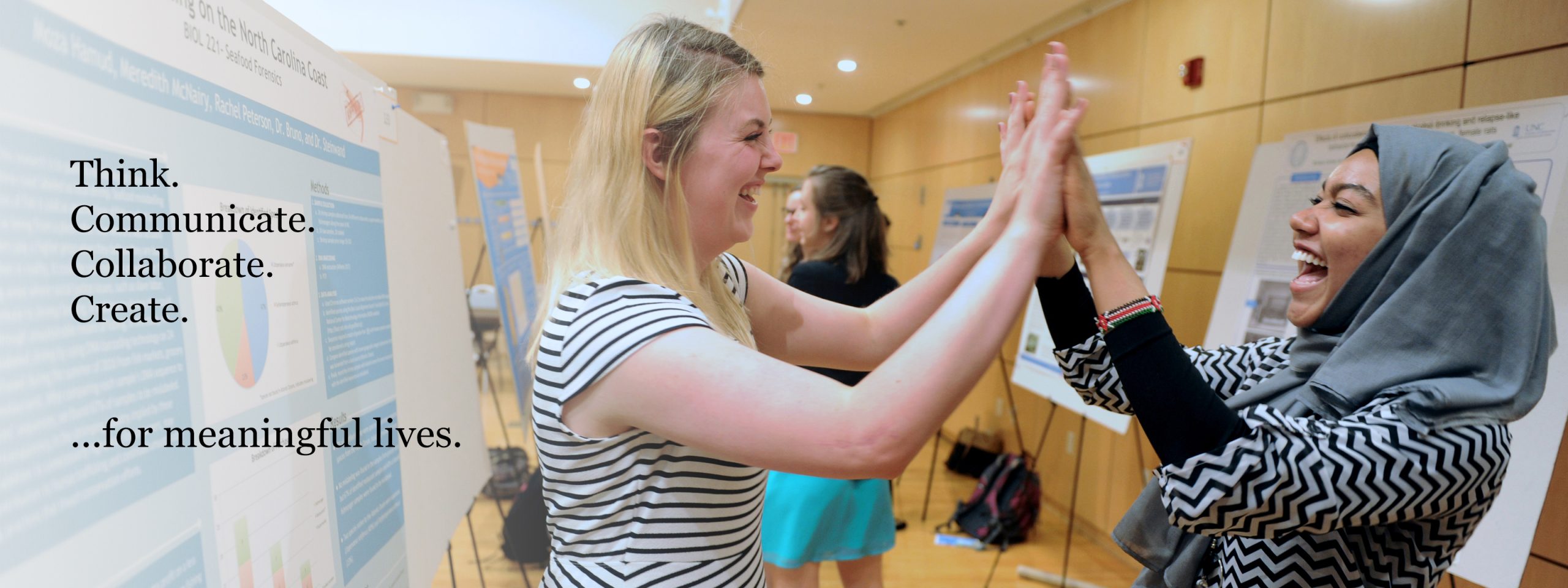 Two students smile and high-five each other at the Celebration of Undergraduate Research. A On a poster to the right, “Think. Communicate. Collaborate. Create. …for meaningful lives.”