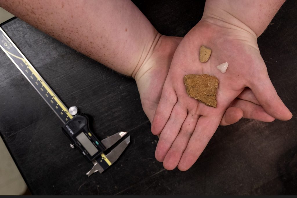 Three small fragments of pottery and stone sit in the palm of Mark Kate's hand. Next to her hand is a metal clamp device to measure the artifacts.