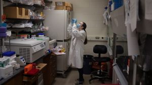 A woman in a lab coat stands in a lab wearing a mask holding up a beaker.