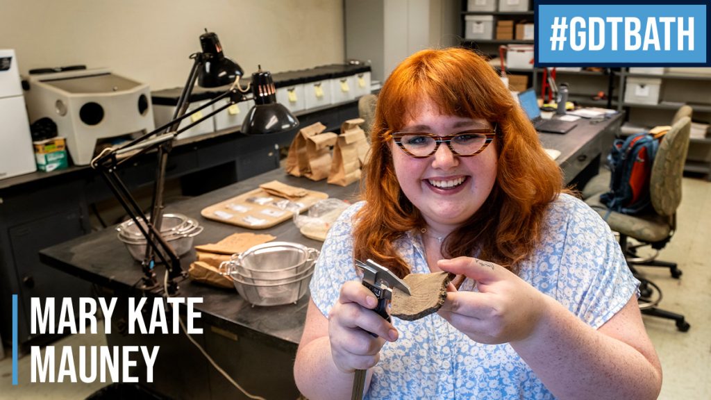 Mary Kate Mauny holds a small piece of pottery in metal clamps. She stands in a lab where artifacts are spread out on a table behind her.