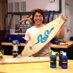 Levi Tox holds a skateboard, which is about three feet in length and features a blue snake design. On the table in front of him is another skateboard he created, a sander and three bottles of paint.