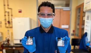 Pedro holds samples of his work in the lab, dressed in lab coast and mask and goggles.