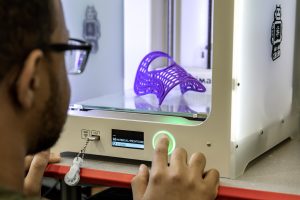 A UNC student looks into a 3D printer. Inside the printer, a purple cylindrical object made of filament has just been printed.