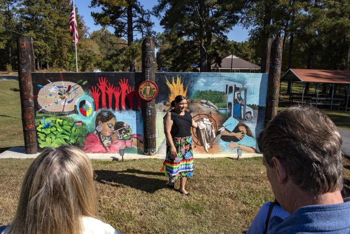 Qua Lynch-Atkins, standing outside, gestures to the mural behind her.