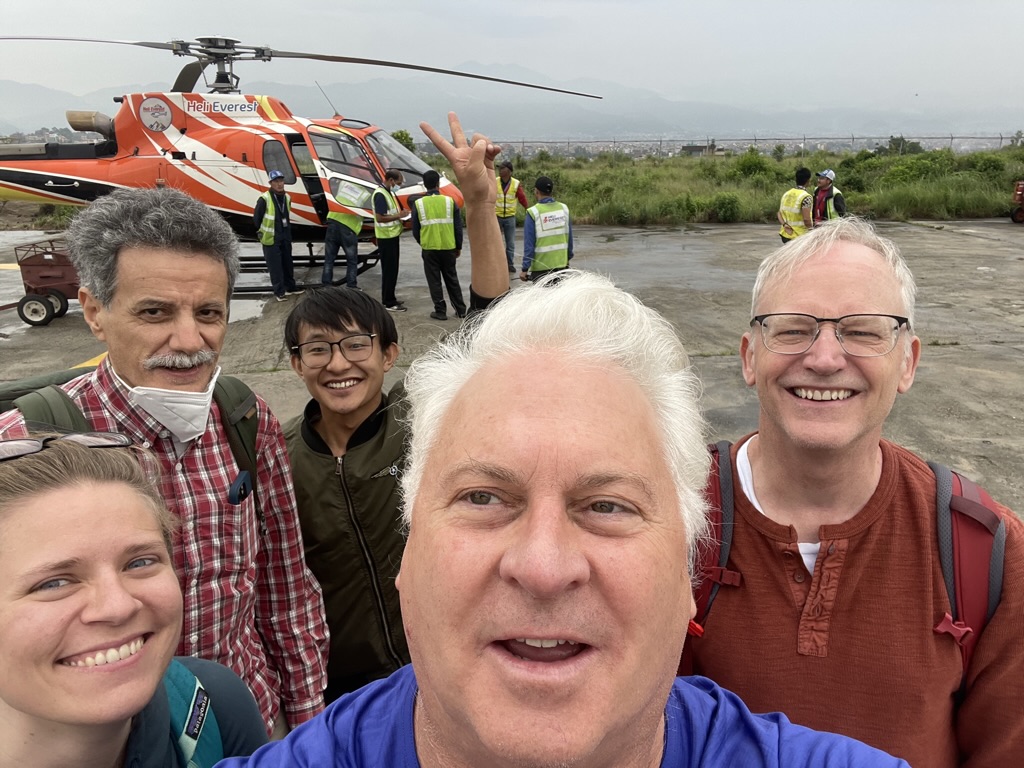 The Nepal summer 2022 team from left to right (with their guide waving in the background): Emily Eidam, Roberto Camassa, Rich McLaughlin and Harvey Seim. They are standing in front of a helicopter.