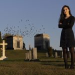 Michelle Freeman stands in a cemetery as birds fly into the sky behind her.