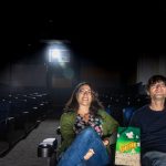 Sabine Gruffat and Bill Brown sit in the front row of a movie theater, a bag of popcorn between them.