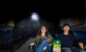 Sabine Gruffat and Bill Brown sit in the front row of a movie theater, a bag of popcorn between them.