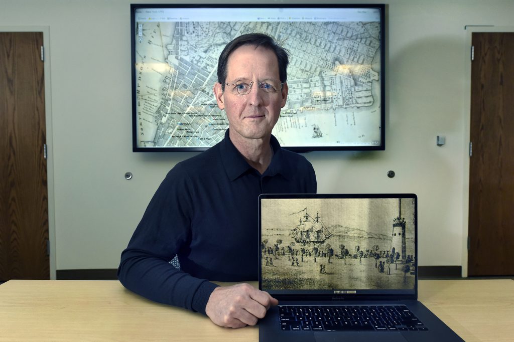 John Sweet sits in front of big screen with a map on it and his laptop with a picture on it related to the book.