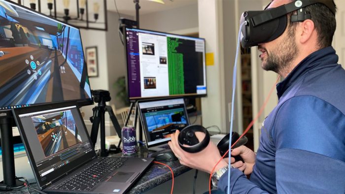 A person wearing a VR headset faces four computer screens with their VR controllers in hand.