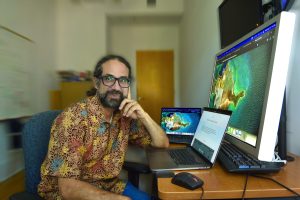 Javier Arce-Nazario sits at his desk with computer screens showing brightly colored maps.