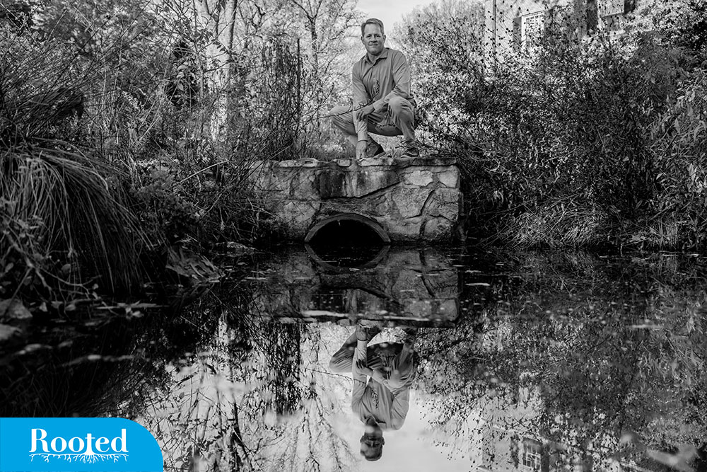 A black and white photo of Mike Piehler sitting on the banks of a creek surrounded by old trees and tree roots and his reflection in the water.
