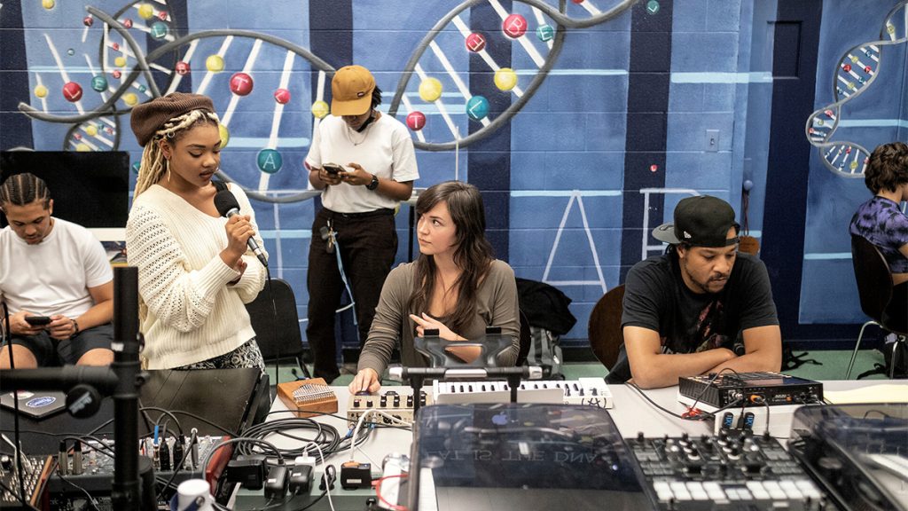 Tirah Phillips sings into a microphone while Katrina Bond and Marc Durham lean over recording equipment. Three other Hip Hop Ensemble members are in the background. 