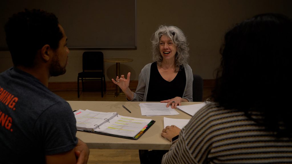 Gwendolyn Schwinke sits at a desk instructing two actors seating across from her.