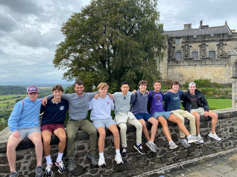 Jackson Holmes and eight other study abroad students sit on a stone wall in front of Stirling Castle.