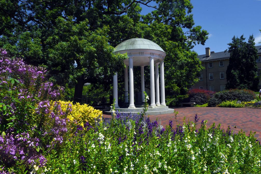 The Old Well in spring, with the foreground filled with purple, yellow and white flowers and the top left filled with a tree. South Building's brick exterior in the background.