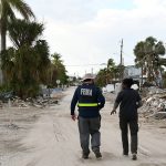 Two FEMA members, one wearing a vest with "FEMA" on the back, on a road lined with rubble and bent trees.