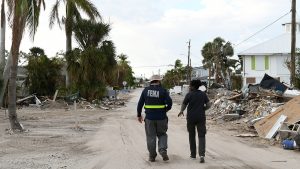 Two FEMA members, one wearing a vest with "FEMA" on the back, on a road lined with rubble and bent trees.
