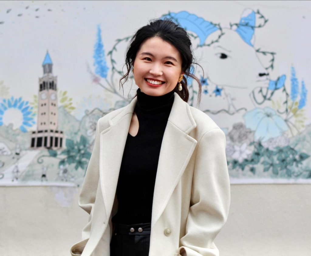 A headshot of Jieni Zhou in front of a mural featuring Rameses the Ram and other UNC landmarks.
