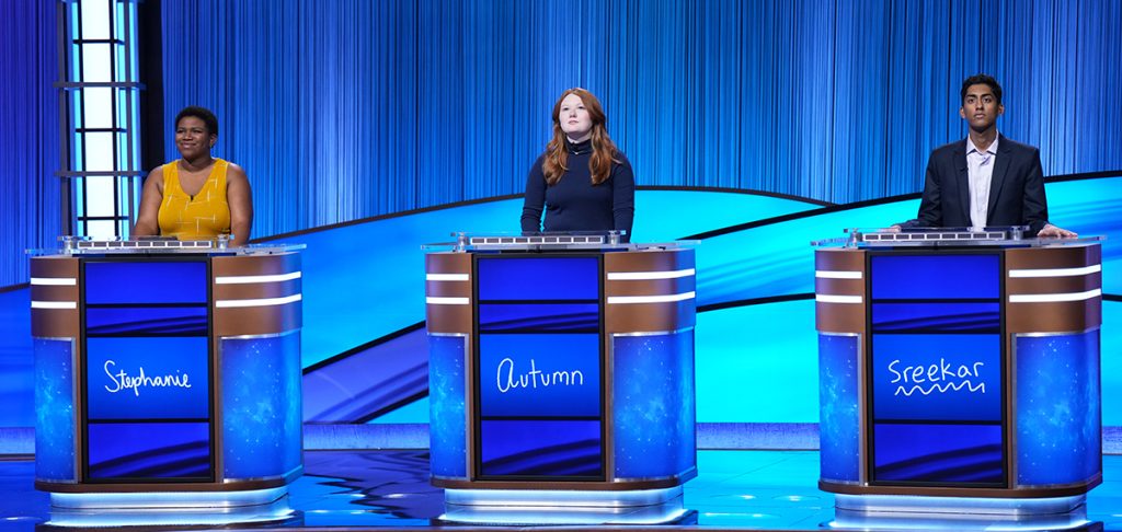 Stephanie Pierson (left) and two other contestants (l-r, Autumn, Sreekar) on the "Jeopardy!" stage behind podiums bearing their names.