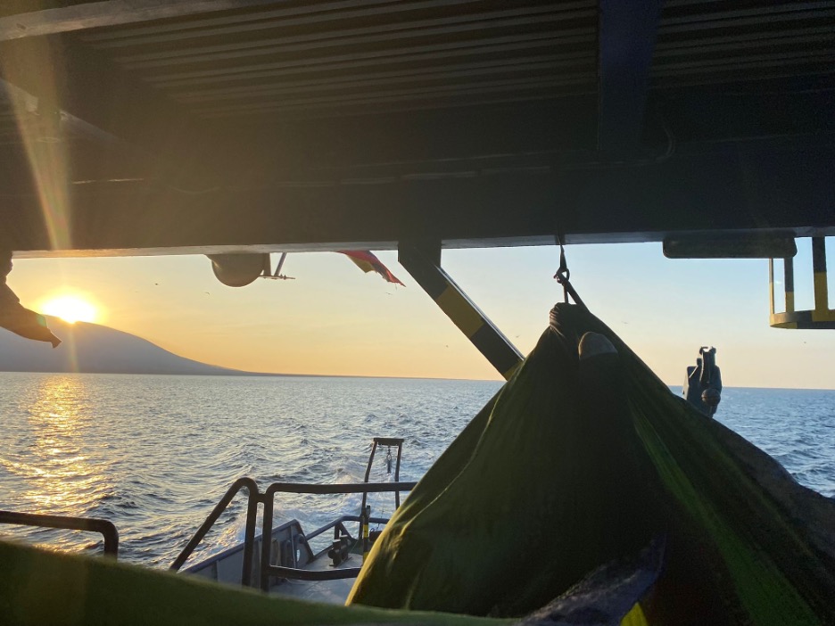 A view of the ocean from the boat with the sun setting over a mountain in the top left corner. The right half shows part of a green hammock.