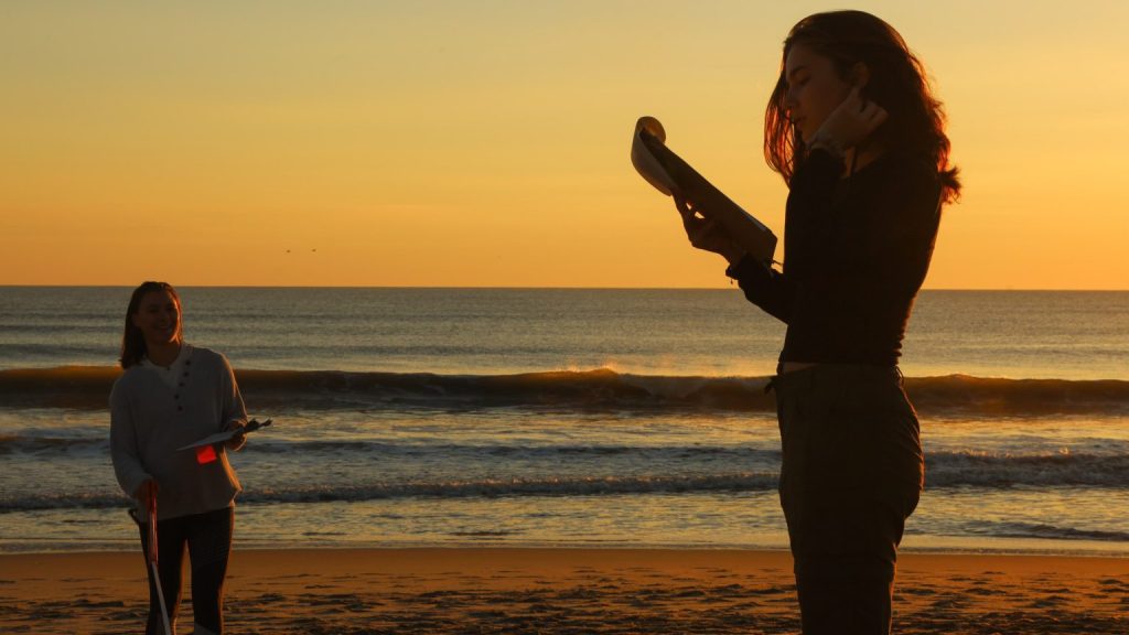 Students Julia Elliott and Anna Smith hold clipboards on the beach at sunrise. 