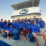 Group photo of the research team wearing matching blue windbreakers, on the front of the boat.