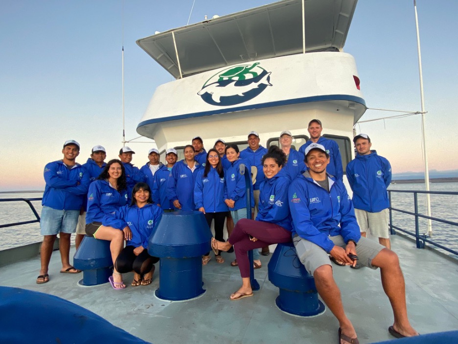 Group photo of the research team wearing matching blue windbreakers, on the deck of the boat.