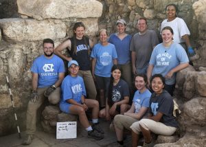 Group photo of the Huqoq team standing at the dig site wearing Carolina attire.