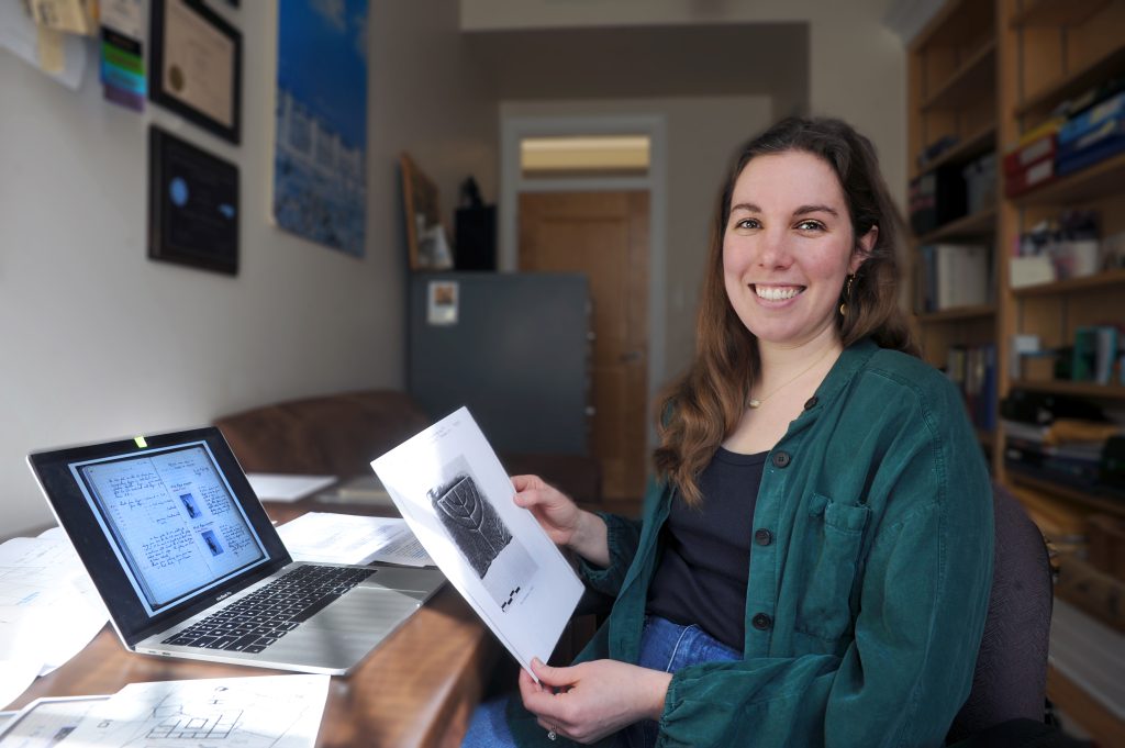 Jocelyn Burney sits at a desk in an office in front of a laptop, holding an archaeological drawing.