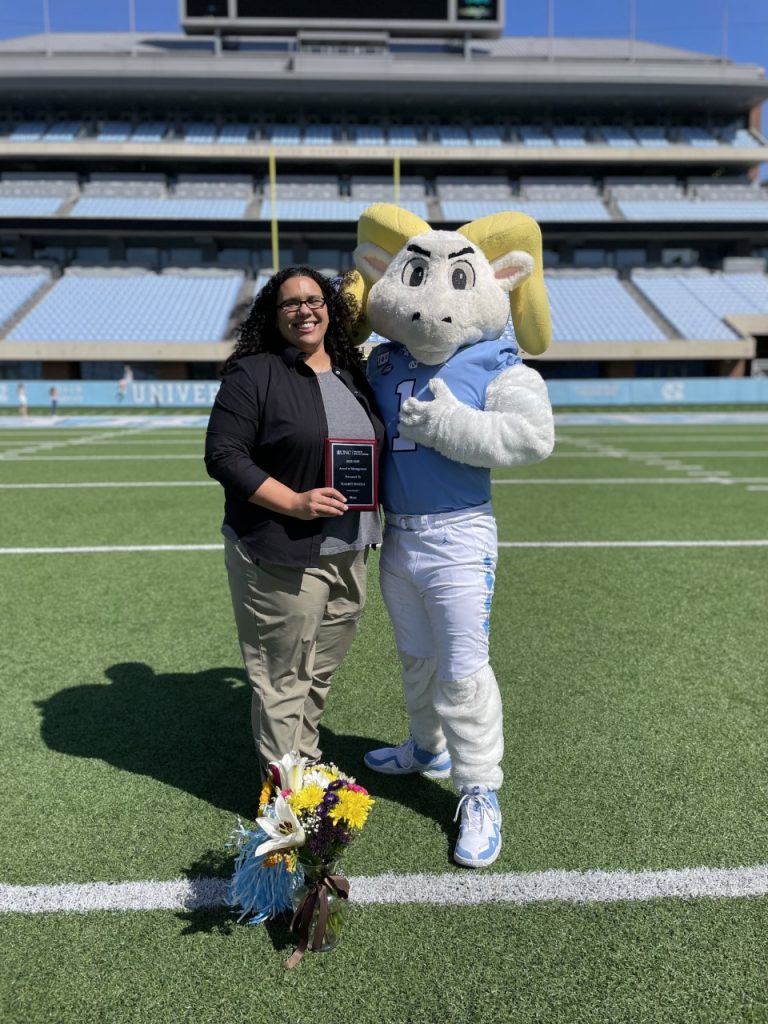 El Fisseha, holding up a plaque, poses with UNC mascot Ramses on the Kenan Stadium field, a vase of flowers at her feet. 